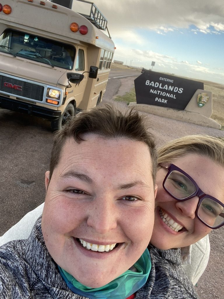 Amanda and Robyn in a selfie at the Badlands National Park entrance sign, with Gus parked behind them.