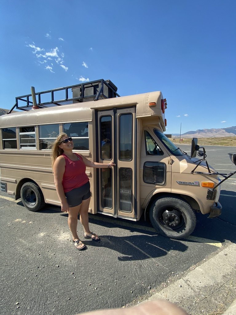 Robyn standing at the open door of Gus, a tan four-window school bus, parked under a blue sky with hills behind.