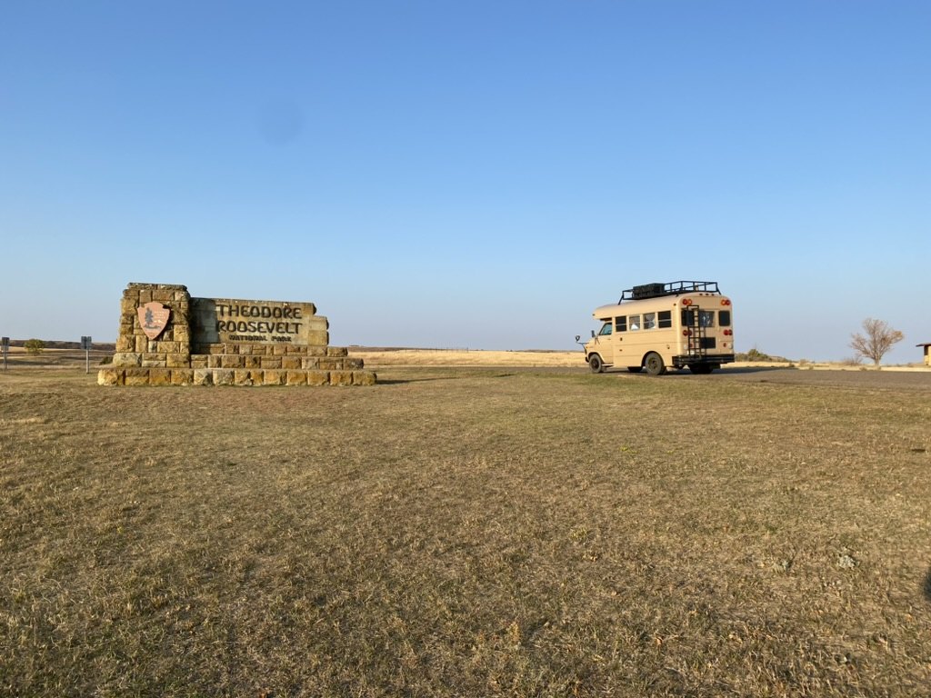 Gus, a tan four-window school bus, parked beside the stone Theodore Roosevelt National Park entrance sign under a clear blue sky.