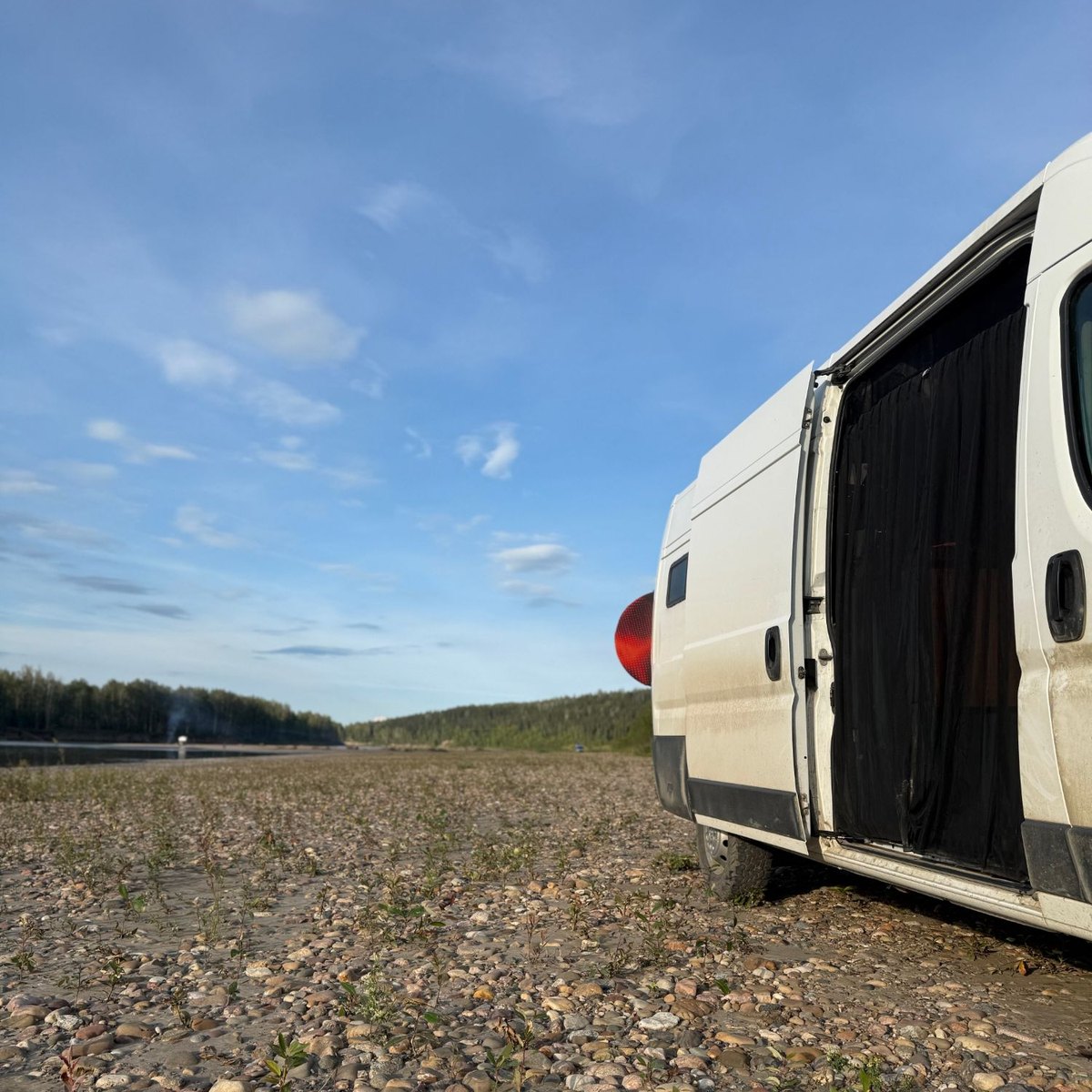 A white camper van parked on a river gravel bar at golden hour, its side door open, forest and water stretching away under a wide sky.
